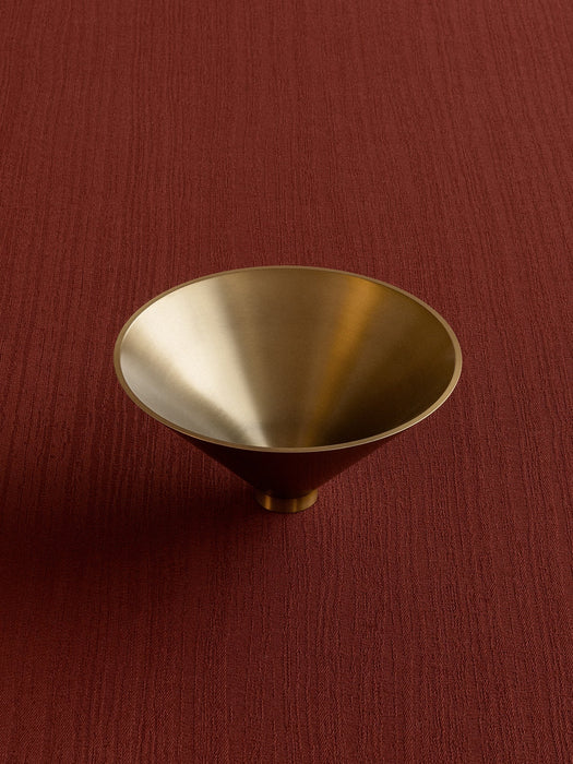 Overhead view of the Audo Taper Bowl in brushed brass. The cone-shaped bowl rests on a textured, deep red surface.