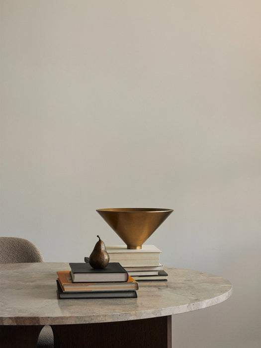 A brass Taper Bowl and a bronze pear-shaped Sentiment Paper Weight are styled with books on a marble Androgyne Dining Table.