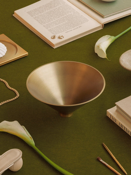 The Audo Taper Bowl in brushed brass on a dark green surface, with books, white calla lilies, and gold jewellery.