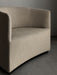 Close-up of the Tearoom Lounge Chair in light beige textured fabric, showing its curved back and armrest against a grey wall