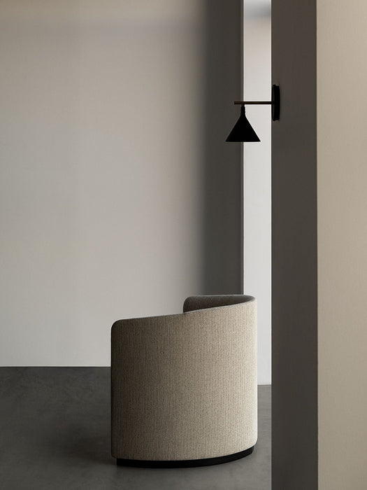 Side view of the beige Tearoom Lounge Chair in a minimalist space with a dark floor next to a black Cast Sconce Wall Lamp.