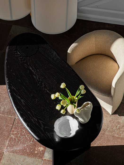 High-angle view of a beige bouclé Tearoom Lounge Chair beside a black coffee table with flowers and ceramics on a tile floor.