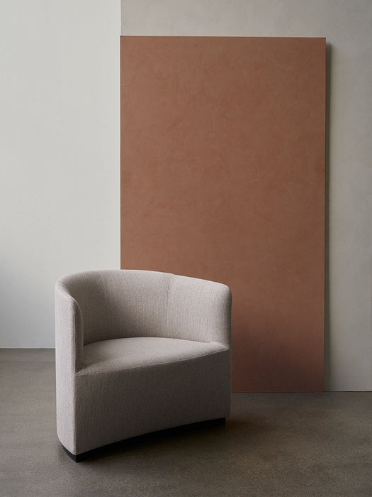 The Tearoom Lounge Chair in light grey upholstery, shown in a minimalist room with a concrete floor and a terracotta panel.