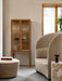 A beige Tearoom Lounge Chair next to a wooden Curiosity Cabinet, with a Cairn Pouf and a Column Table Lamp on a marble Plint.