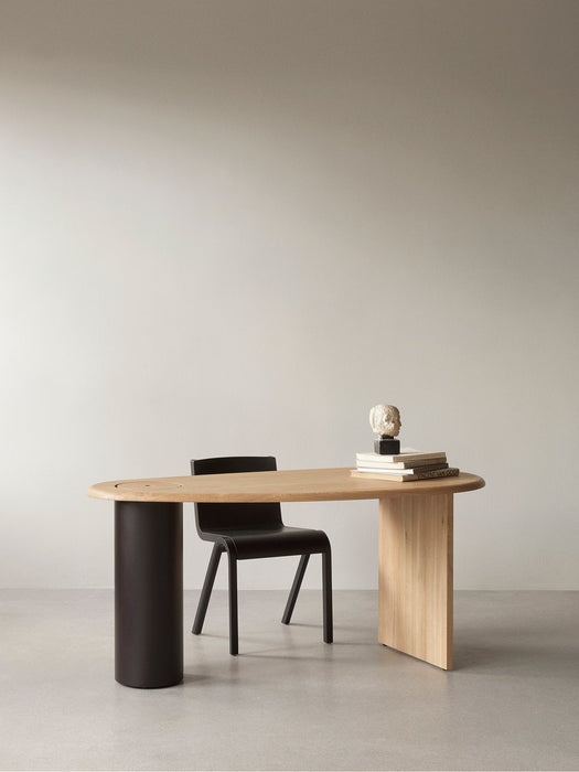 The asymmetrical Eclipse Desk in natural oak, styled with a black chair. On the desk sits a stack of books and a sculpture.
