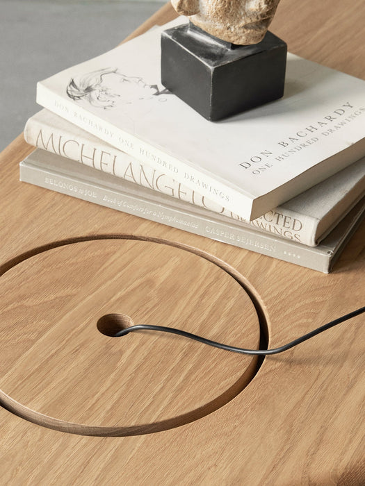 A close-up of The Eclipse Desk in natural oak, showing the circular storage lid with a cable, books and a small sculpture.