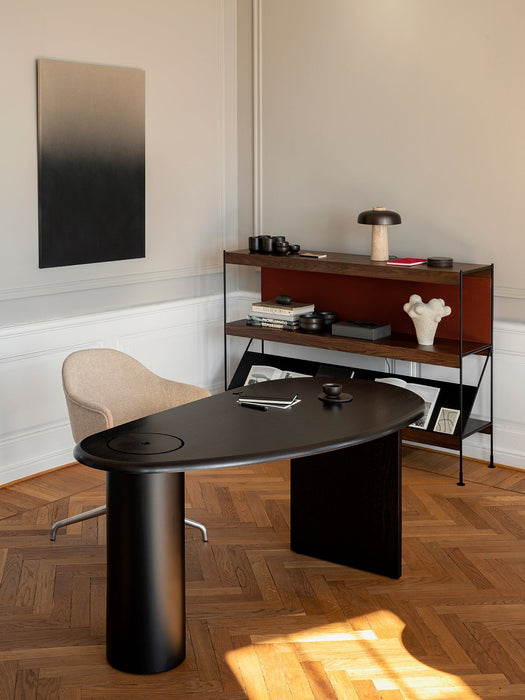 The black lacquered oak Eclipse Desk in a modern office with a beige chair, wooden shelving unit, and abstract wall art.