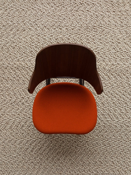 A top-down view of The Penguin Dining Chair, with a walnut backrest and orange upholstered seat, on a textured beige rug.