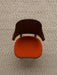 A top-down view of The Penguin Dining Chair, with a walnut backrest and orange upholstered seat, on a textured beige rug.