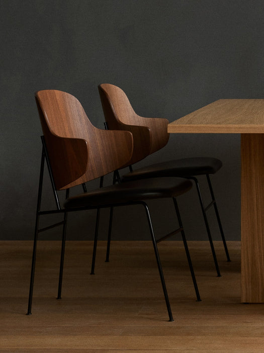 Two Penguin Dining Chairs in walnut with black seats tucked under an oak Androgyne Dining Table against a dark grey wall.