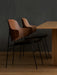 Two Penguin Dining Chairs in walnut with black seats tucked under an oak Androgyne Dining Table against a dark grey wall.