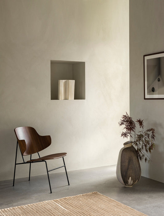 The Penguin Lounge Chair, with its dark wood seat and black frame, sits in a serene, minimalist room next to a large vase.