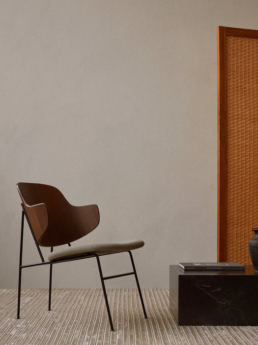 The Penguin Lounge Chair with a walnut back and light seat, placed on a striped rug next to a black marble plinth table.