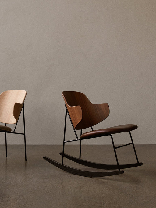 The Penguin Rocking Chair in walnut with a leather seat stands in a minimal room next to The Penguin Lounge Chair in oak.