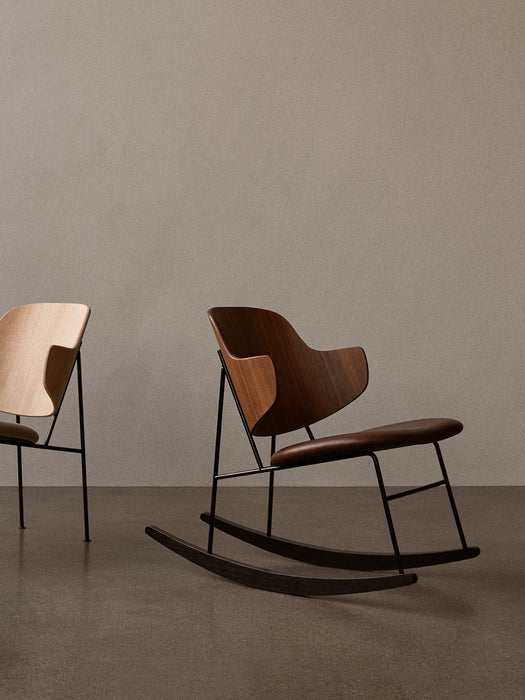 The Penguin Rocking Chair in walnut with a leather seat stands in a minimal room next to The Penguin Lounge Chair in oak.