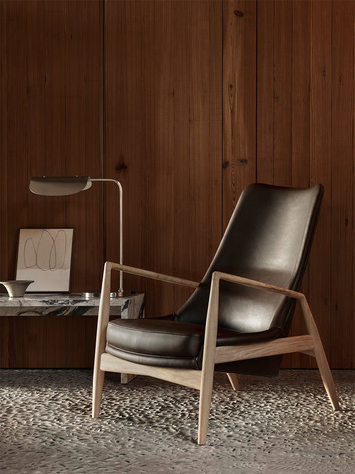 The Seal Lounge Chair, High Back in dark leather with an oak frame, styled in a room with wood walls next to a marble table.