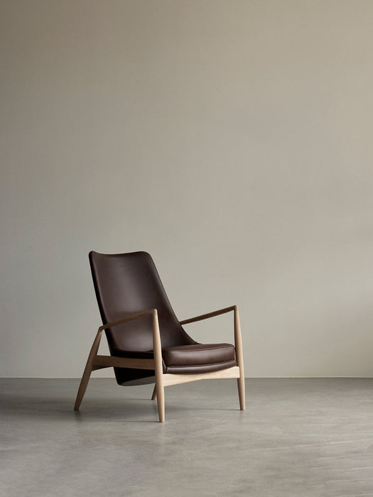 The Seal Lounge Chair, High Back, in brown leather with a light wood frame, sits in an empty room with grey floors and walls.