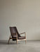 The Seal Lounge Chair, High Back, in brown leather with a light wood frame, sits in an empty room with grey floors and walls.