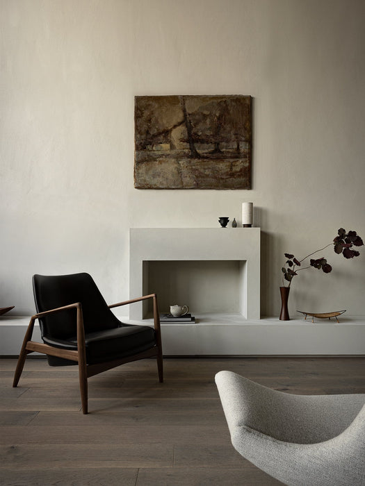 Side view of The Seal Lounge Chair in black leather and dark wood, styled in a room with a fireplace and a grey Pagode Sofa.