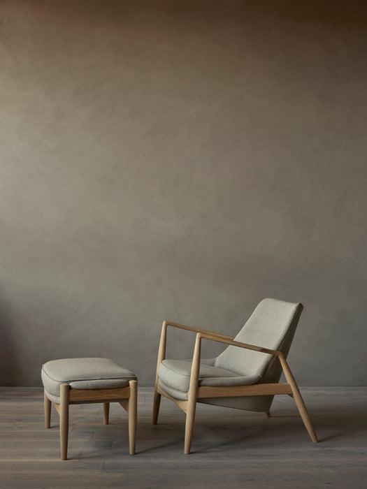 A side view of The Seal Lounge Chair and Seal Ottoman with natural oak frames and grey upholstery, on a dark wood floor.