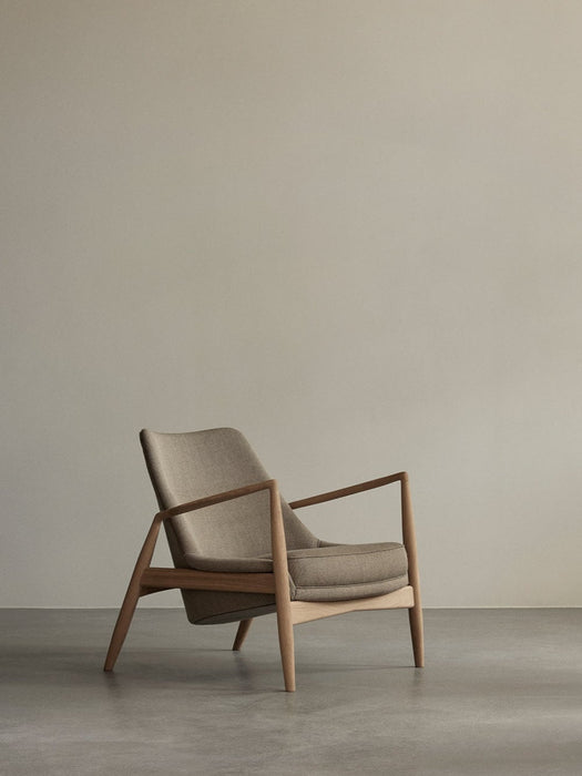 Side-angle view of The Seal Lounge Chair with a low back, light wood frame and beige fabric upholstery, on a grey floor.