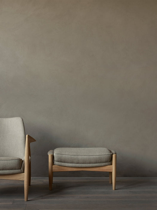 The Seal Ottoman in grey fabric and oak sits next to the Seal Lounge Chair on a dark wood floor against a taupe wall.