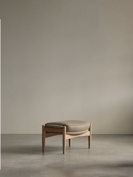 The Seal Ottoman with a natural oak frame and a thick, light brown cushion, shown in a minimalist room on a concrete floor.