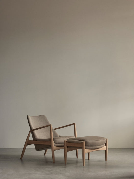 The Seal Lounge Chair and The Seal Ottoman, with light wood frames and beige fabric, in a minimalist room with grey floors.