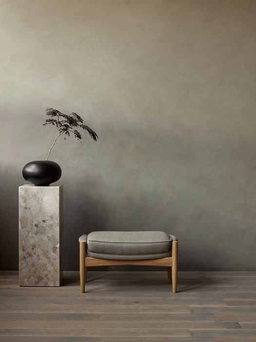The Seal Ottoman with a grey upholstered cushion sits on a wood floor next to a marble Plinth Pedestal holding a black vase.