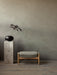 The Seal Ottoman with a grey upholstered cushion sits on a wood floor next to a marble Plinth Pedestal holding a black vase.