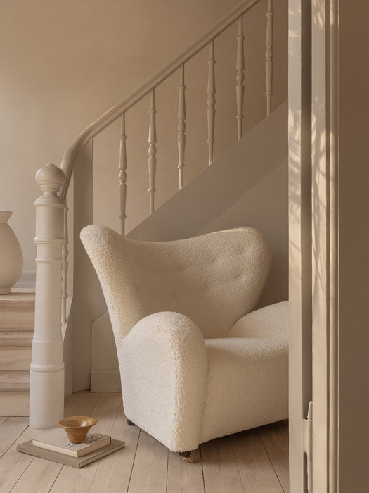 The Tired Man Lounge Chair in white boucle sits under a staircase. A Triptych Bowl rests on books on the light wood floor.