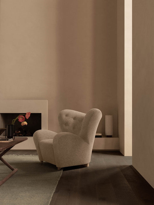 A beige Tired Man Lounge Chair in a minimalist room with a fireplace, coffee table, Kubus Vase, and a Hahira Lamp.