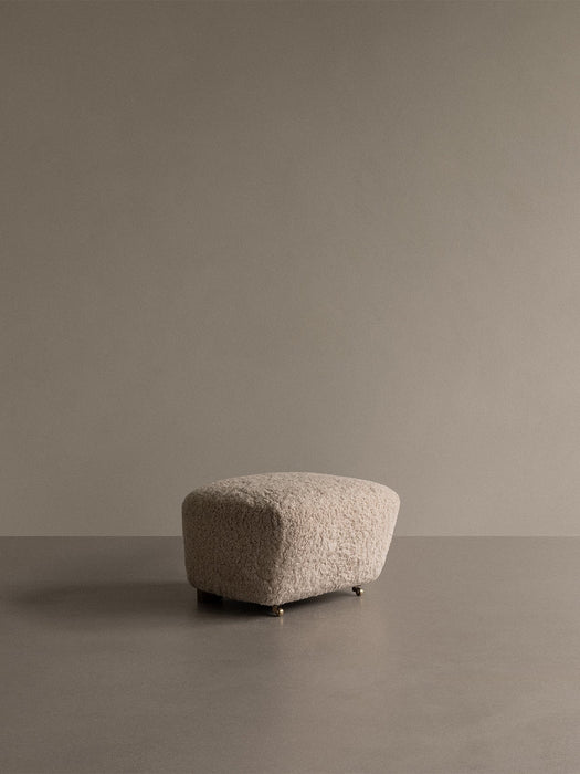 The Tired Man Ottoman, upholstered in light sheepskin, shown in a minimalist room with a taupe wall and floor.
