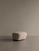 The Tired Man Ottoman, upholstered in light sheepskin, shown in a minimalist room with a taupe wall and floor.