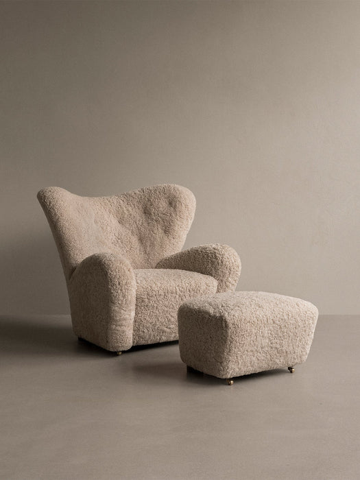 Light sheepskin upholstered Tired Man Armchair and matching Tired Man Ottoman with brass castors, shown in a studio setting.