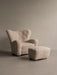 Light sheepskin upholstered Tired Man Armchair and matching Tired Man Ottoman with brass castors, shown in a studio setting.