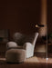 A Tired Man Lounge Chair and ottoman in soft, off-white sheepskin styled in a dark room next to an illuminated floor lamp.