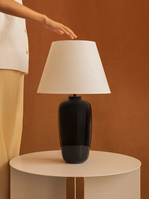 A hand hovers over the white shade of a black Torso Table Lamp on a beige Androgyne Side Table against a brown wall.