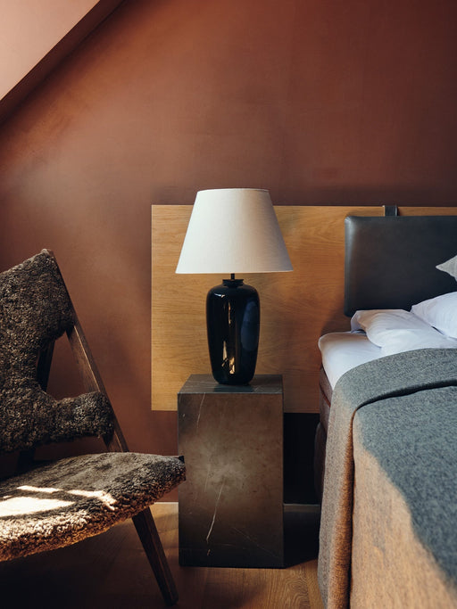 The black Torso Table Lamp, 57 on a marble plinth as a bedside lamp in a bedroom with a sheepskin chair and terracotta walls.
