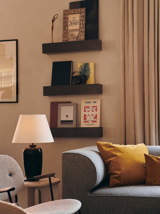 A warmly lit Torso Table Lamp, 57 on a side table in a living room setting, next to a grey sofa and dark decorative shelves.