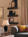 A warmly lit Torso Table Lamp, 57 on a side table in a living room setting, next to a grey sofa and dark decorative shelves.