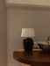 The black ceramic Torso Portable Table Lamp with a linen shade sits on a dark wood Androgyne Side Table in a softly lit room.
