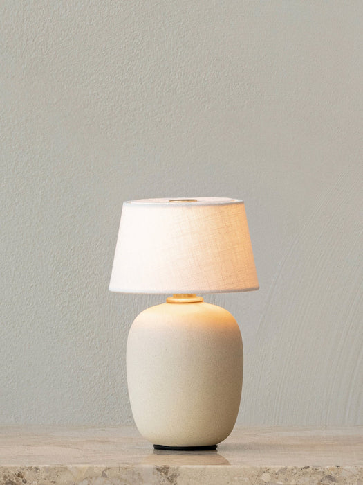 The Torso Portable Table Lamp in Sand, illuminated and standing on a marble surface against a plain, light grey wall.