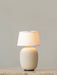 The Torso Portable Table Lamp in Sand, illuminated and standing on a marble surface against a plain, light grey wall.