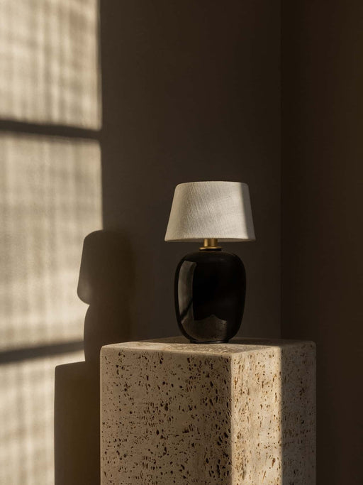 The portable Torso Table Lamp with a black ceramic base and linen shade sits on a travertine plinth in a sunlit corner.