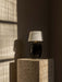 The portable Torso Table Lamp with a black ceramic base and linen shade sits on a travertine plinth in a sunlit corner.