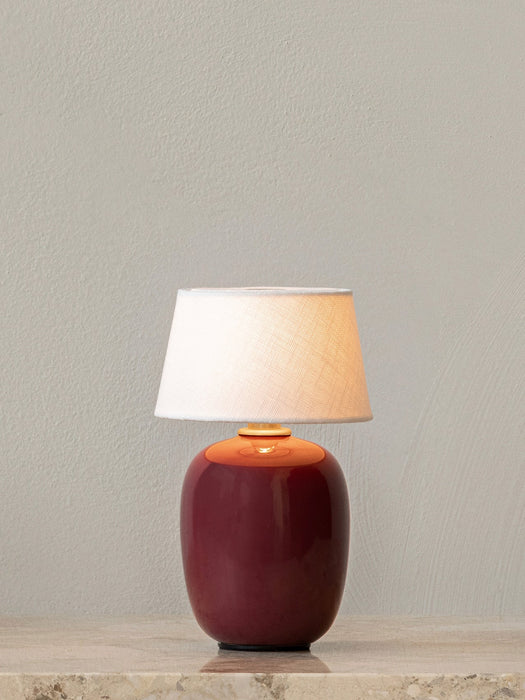 The Torso Portable Table Lamp in Ruby, with its glowing linen shade, sitting on a marble tabletop against a light grey wall.