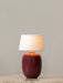 The Torso Portable Table Lamp in Ruby, with its glowing linen shade, sitting on a marble tabletop against a light grey wall.