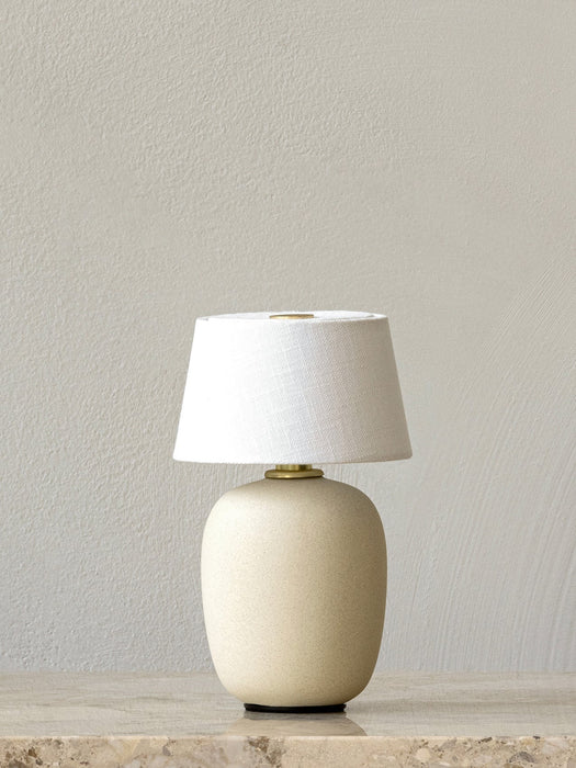 The Sand Torso Portable Table Lamp with a ceramic base and white linen shade sits on a marble surface against a plain wall.