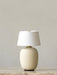 The Sand Torso Portable Table Lamp with a ceramic base and white linen shade sits on a marble surface against a plain wall.